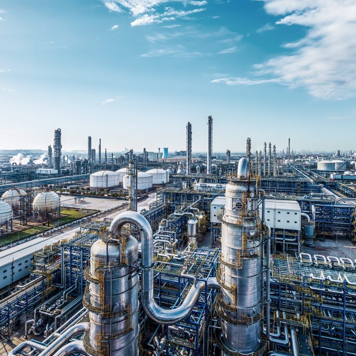 Große Industrieanlage mit Türmen, Tanks und verbundenen Rohrleitungen unter einem strahlend blauen Himmel, mit Grünflächen im Hintergrund.