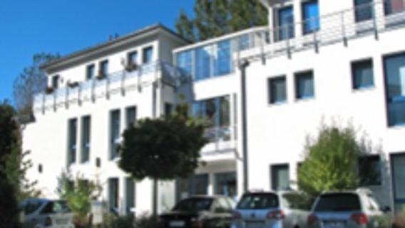 A white building with multiple floors, surrounded by greenery and parked cars.